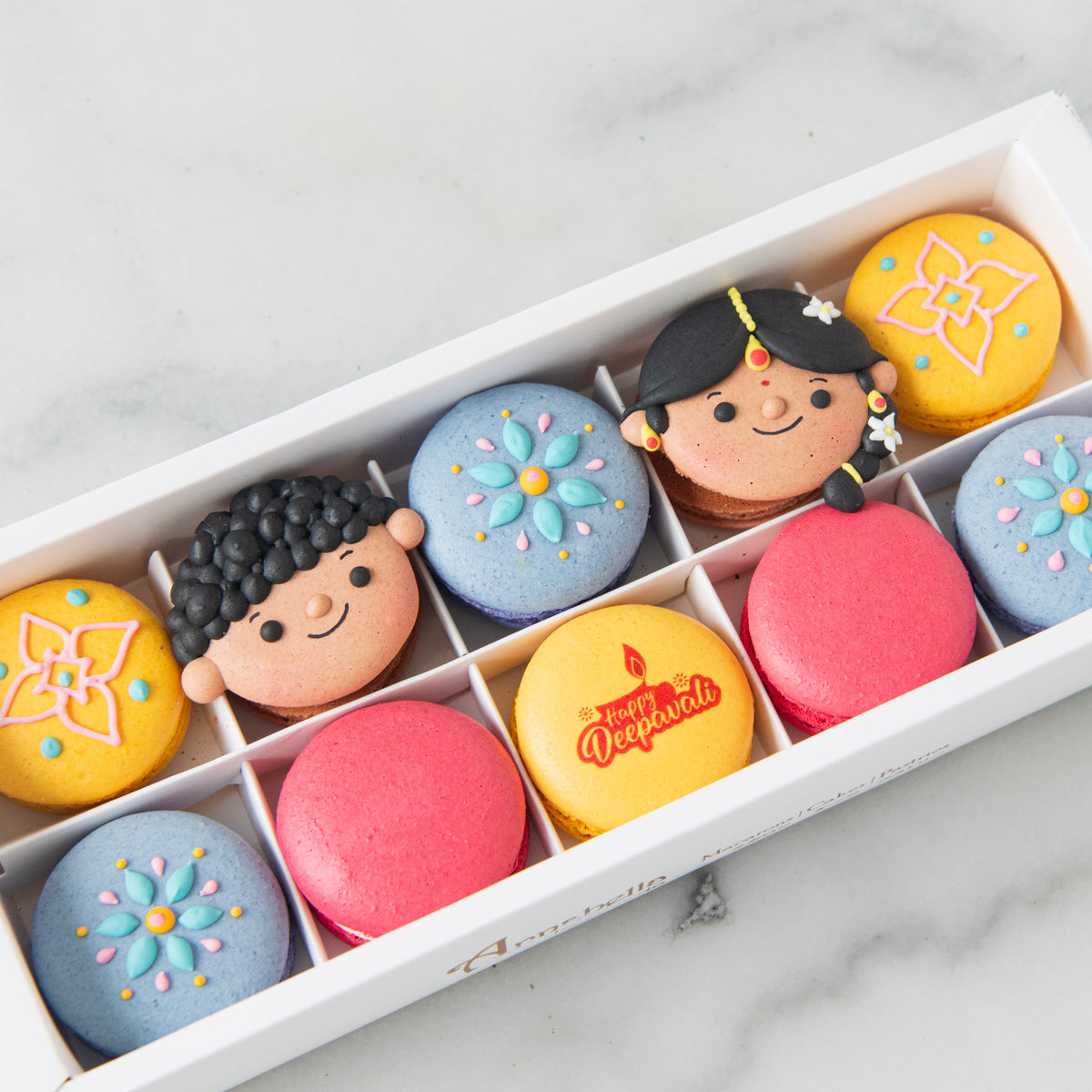 A white gift box filled with decorated macarons on a light marble surface. The macarons include pastel blue, pink, and yellow flavors, with some decorated to look like cartoon characters and floral motifs.