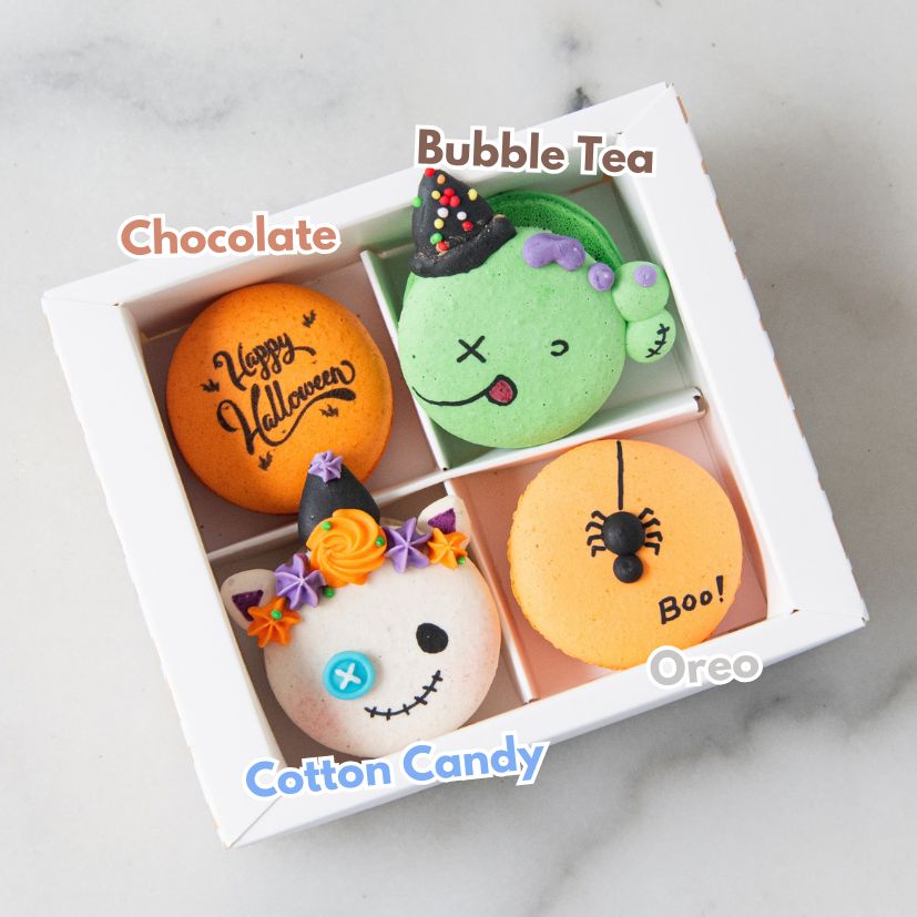 A white gift box containing four Halloween-themed macarons labeled Chocolate, Bubble Tea, Cotton Candy, and Oreo, arranged on a light marble surface.