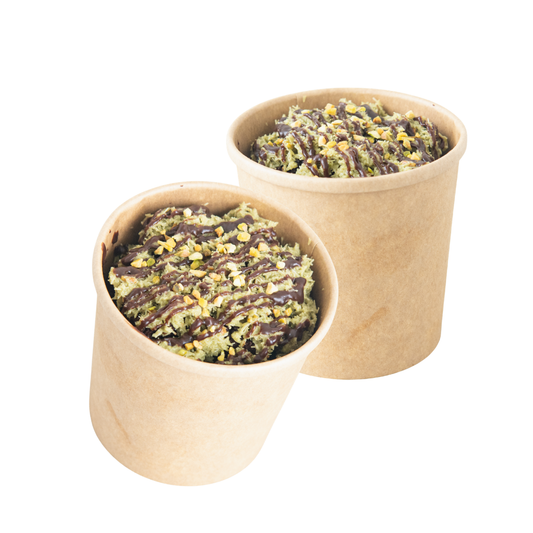 Two paper cup desserts filled with a green pistachio-like mixture and drizzled with chocolate sauce, shown on a plain white background.