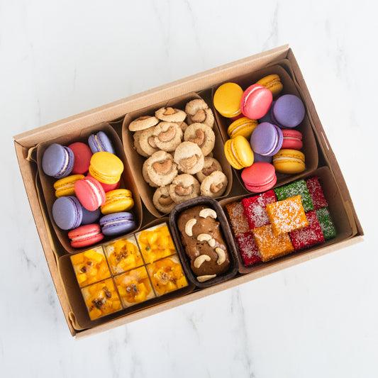 A top-down view of a brown cardboard gift box filled with assorted sweets and snacks, including colorful macarons, cookies, small cakes, and decorated candies, set on a light marble surface.