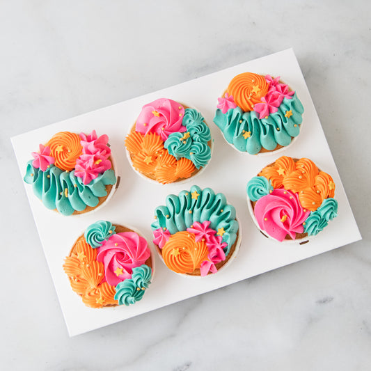 Six decorated cupcakes arranged on a white rectangular tray over a light marble surface. The cupcakes have colorful piped frosting in orange, pink, and teal, with small yellow sprinkle accents.