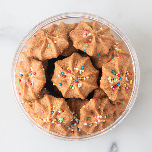 Star-shaped sugar cookies sprinkled with colorful candy melts, presented in a clear round plastic container on a light marble surface.