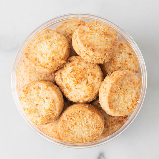 A top-down view of round, golden-brown cookies coated with shredded coconut inside a clear plastic tub, set against a light gray marble background.