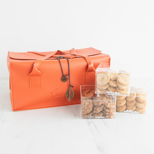 An orange faux-leather gift bag with a flap closure, dual handles, and a hanging bronze leaf charm, displayed alongside three clear plastic containers filled with assorted cookies on a white marble surface against a white background.