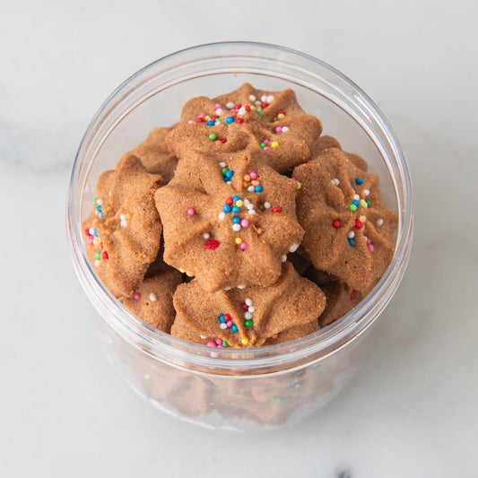 Star-shaped brown cookies topped with colorful sprinkles, displayed in a clear plastic jar on a light marble surface.