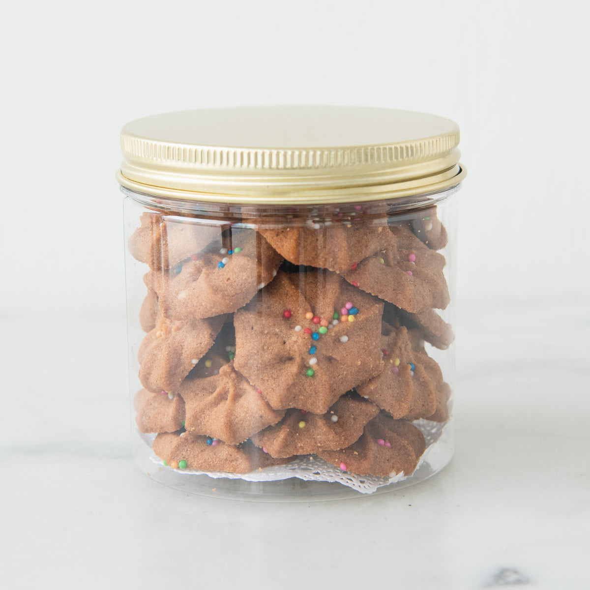 A clear cylindrical glass jar filled with brown star-shaped cookies topped with colorful sprinkles, sealed with a metallic gold lid, on a light gray surface.