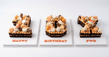 Three custom chocolate and macaron number cakes displayed side-by-side on white stands against a clean white background. The left cake is shaped like the number 8 and is decorated with orange and beige macarons and chocolate, with 'HAPPY' written in orange at the base.